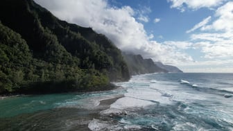 Aerial view along the Na Pali Coast of Kauai, Hawaii, with steep green cliffs dropping into white surf and blue-green reef shallows under a partly cloudy sky.